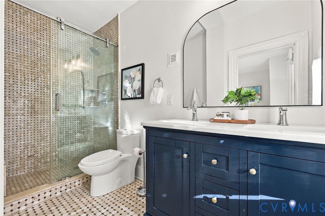 522 N 27th Street, Church Hill, Richmond, VA 23223 Bathroom featuring a double-sink vanity with a solid-surface countertop, an arched wall-mounted mirror, and a walk-in shower with a sliding glass door and mosaic tile surround