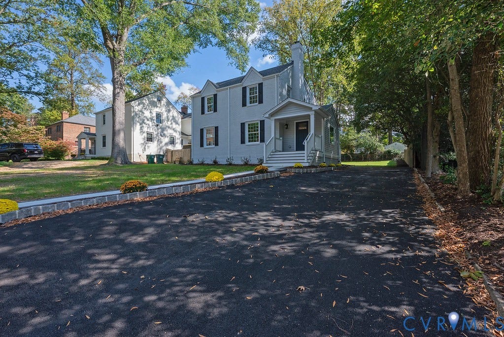 Two-story exterior featuring light-toned siding, black window shutters, a covered front entryway, and an expansive asphalt driveway