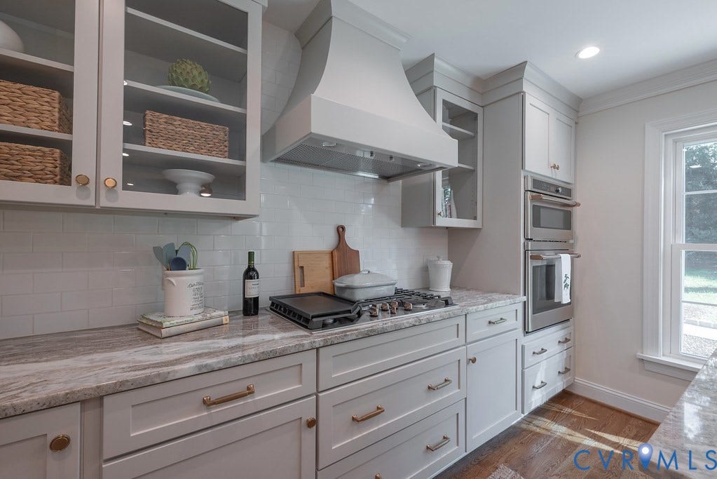 Kitchen featuring custom shaker-style cabinetry, patterned stone countertops, white subway tile backsplash, stainless steel double wall oven, and a built-in range hood