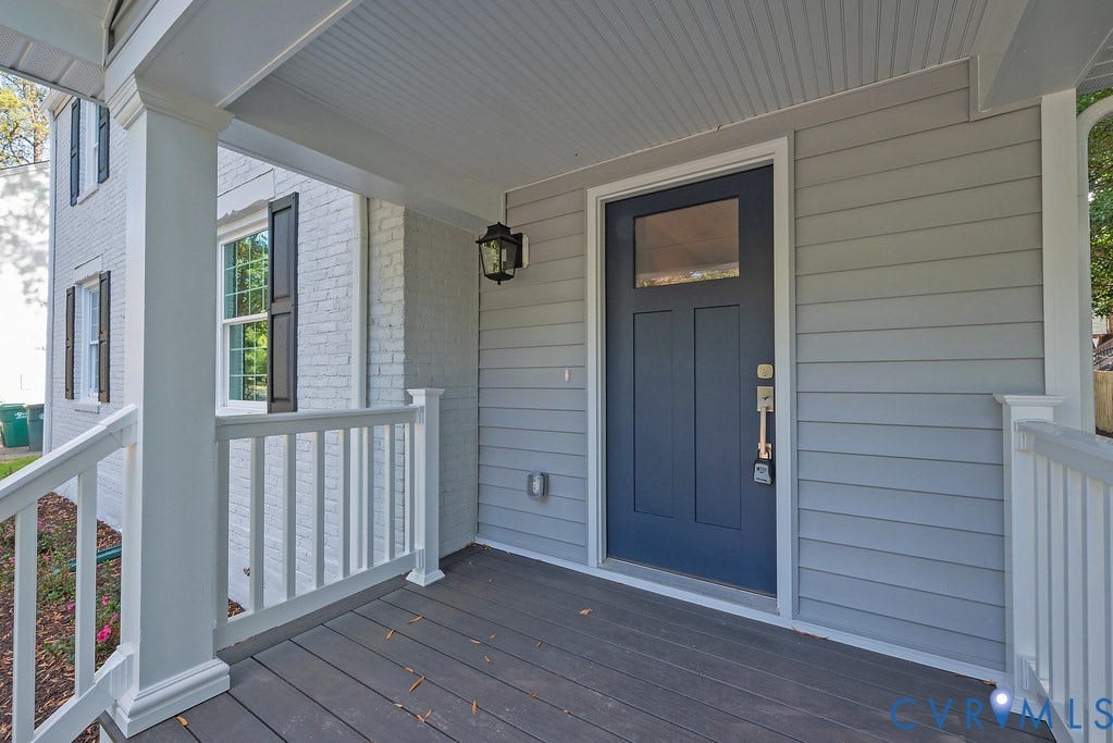 Covered front porch featuring gray wood-finish decking, painted lap siding, a solid-panel door with a single glass pane, white railing, and a wall-mounted lantern light