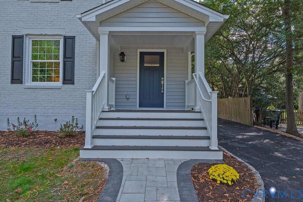 Covered front porch with white railing, blue exterior door, light-toned brick facade, dark window shutters, and a stone-paver walkway