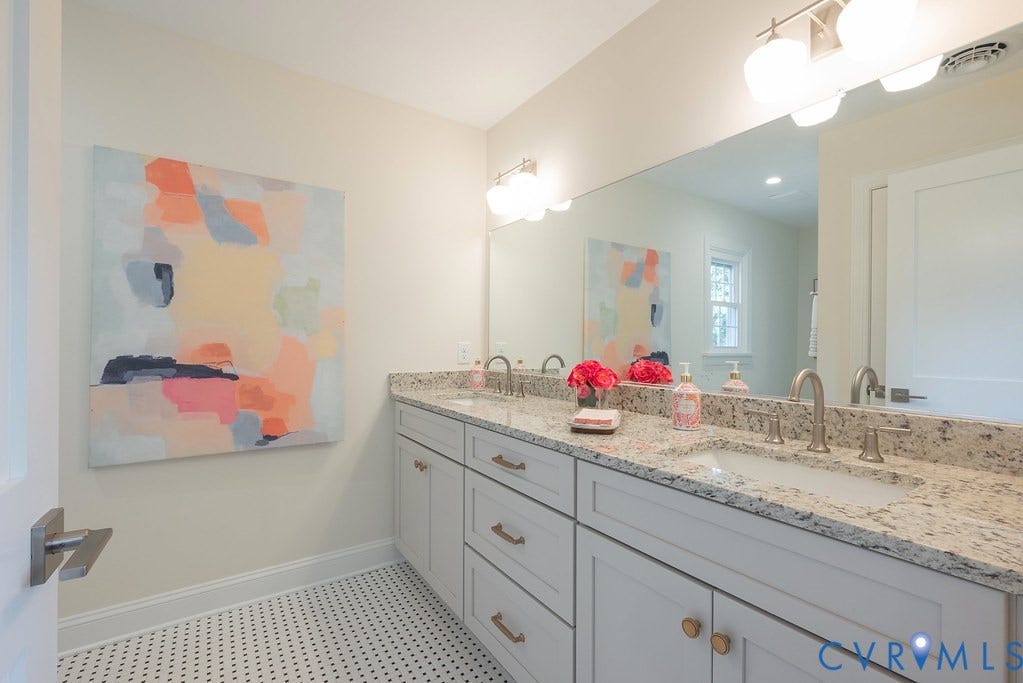 Bathroom featuring a dual-sink vanity with light-toned cabinetry, light-flecked stone countertop, under-mount sinks, brushed metal hardware, and mosaic-style floor tiling