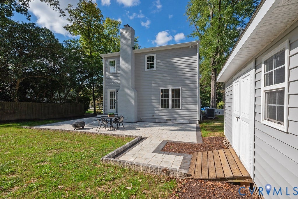 Exterior rear elevation featuring light grey horizontal siding, a painted brick chimney, stone paver patio, and detached storage structure with white paneled doors
