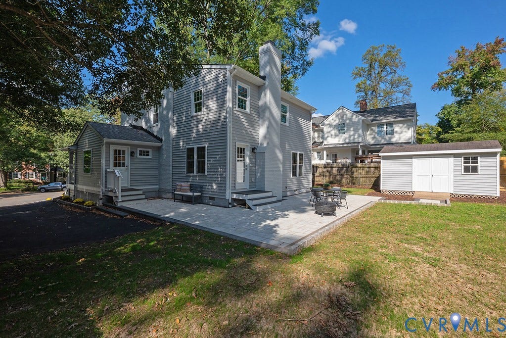 Two-story residence with grey horizontal siding, a light-toned paver patio, external masonry chimney, and a detached matching storage building