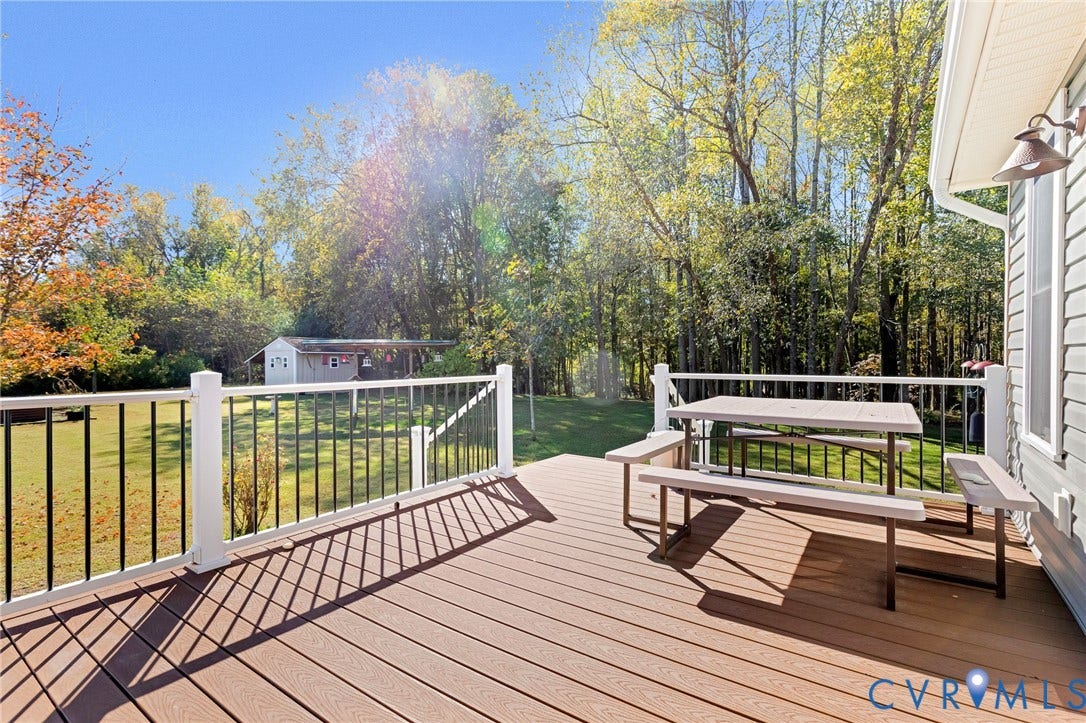 3820 Upshaw Road, Decker's Pond, Aylett, VA 23009 Elevated rear deck featuring composite decking, white posts, black metal balusters, and a wall-mounted exterior light fixture, overlooking a backyard with a detached storage structure and dense tree line