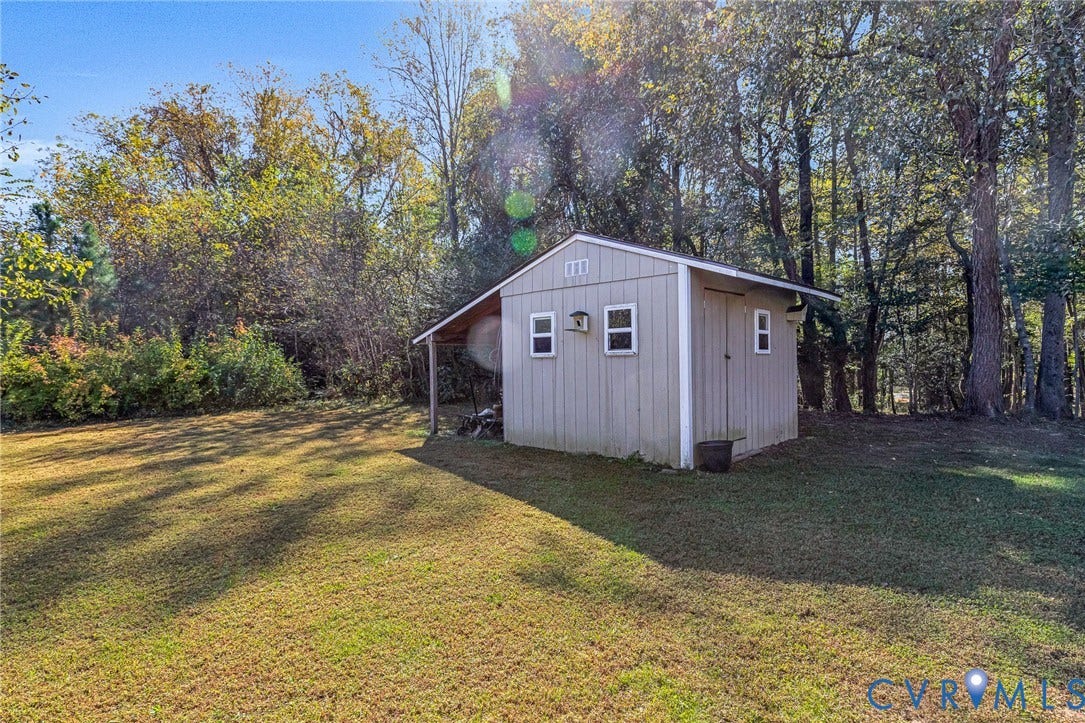 3820 Upshaw Road, Decker's Pond, Aylett, VA 23009 Detached exterior storage structure featuring vertical siding, three windows, and a covered side lean-to, situated on a grass lot with mature perimeter trees