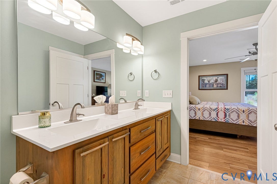 3820 Upshaw Road, Decker's Pond, Aylett, VA 23009 Dual-sink vanity with natural wood-finish cabinetry, solid surface countertop, satin nickel hardware, and large vanity mirror with integrated wall-mounted lighting