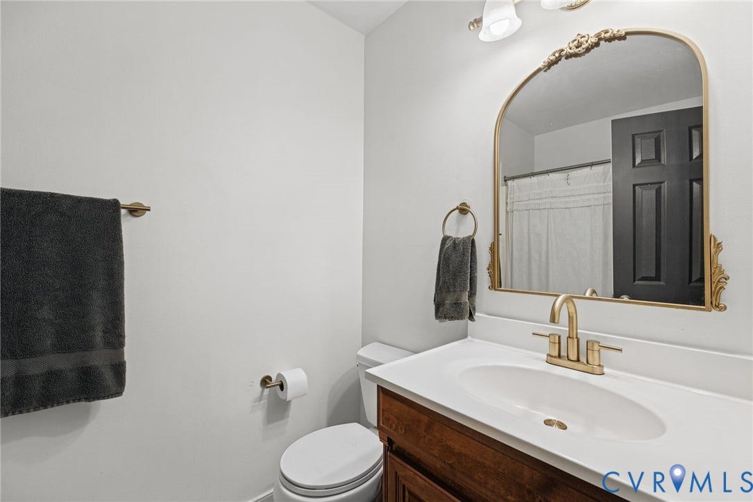 Bathroom featuring a wood-tone vanity, white countertop with integrated oval sink, brass-finish faucet, and an arched wall mirror with decorative gold-tone frame