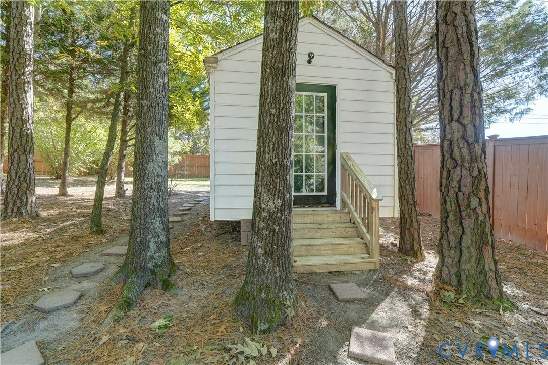 5 Bridgehampton Place, Yorkshire, Henrico, VA 23229 Detached outbuilding featuring white horizontal siding, a glass-paneled entry door, and timber stairs with railings, situated within a wooded lot with stone path pavers and a privacy fence