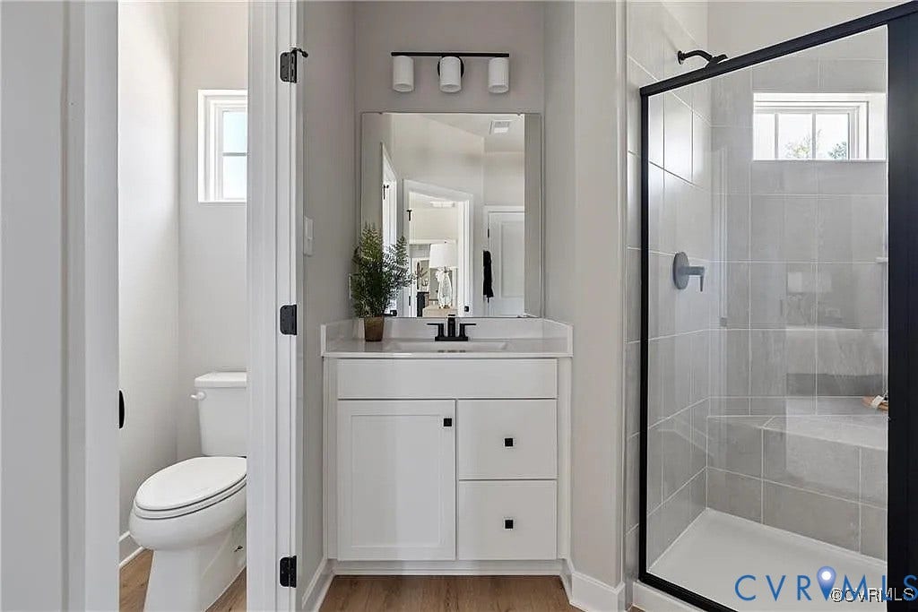 Bathroom featuring a white vanity with matte black hardware, a tiled shower with a glass enclosure, wood-finish flooring, and a three-light vanity fixture