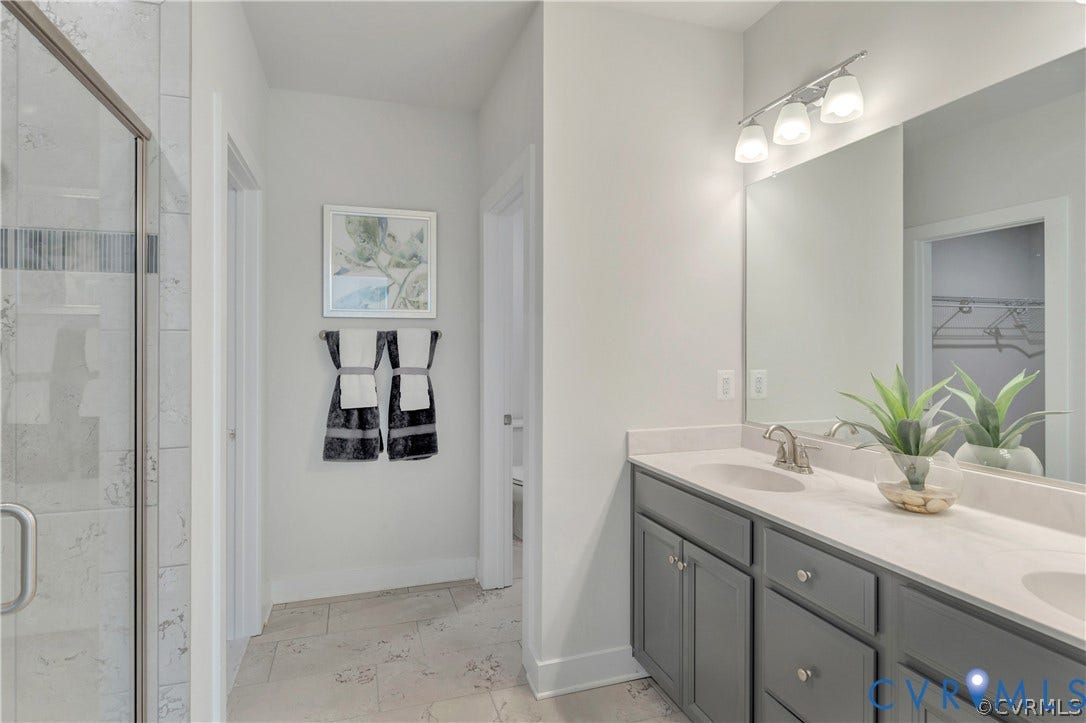 Dual-sink vanity with gray cabinetry and stone-composite countertop, full-width mirror, glass-enclosed shower with tiled surround, and ceramic tile flooring