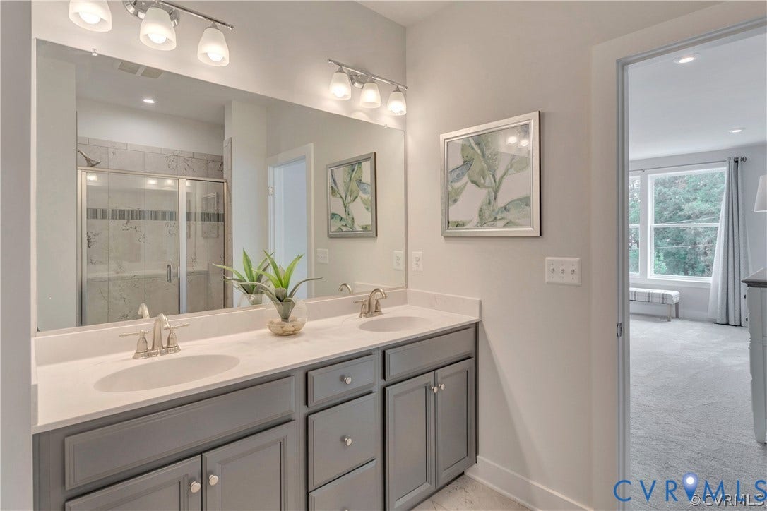 Dual-sink vanity with a solid-surface countertop, gray cabinetry, large plate-glass mirror, polished metal fixtures, and walk-in shower with tiled surround