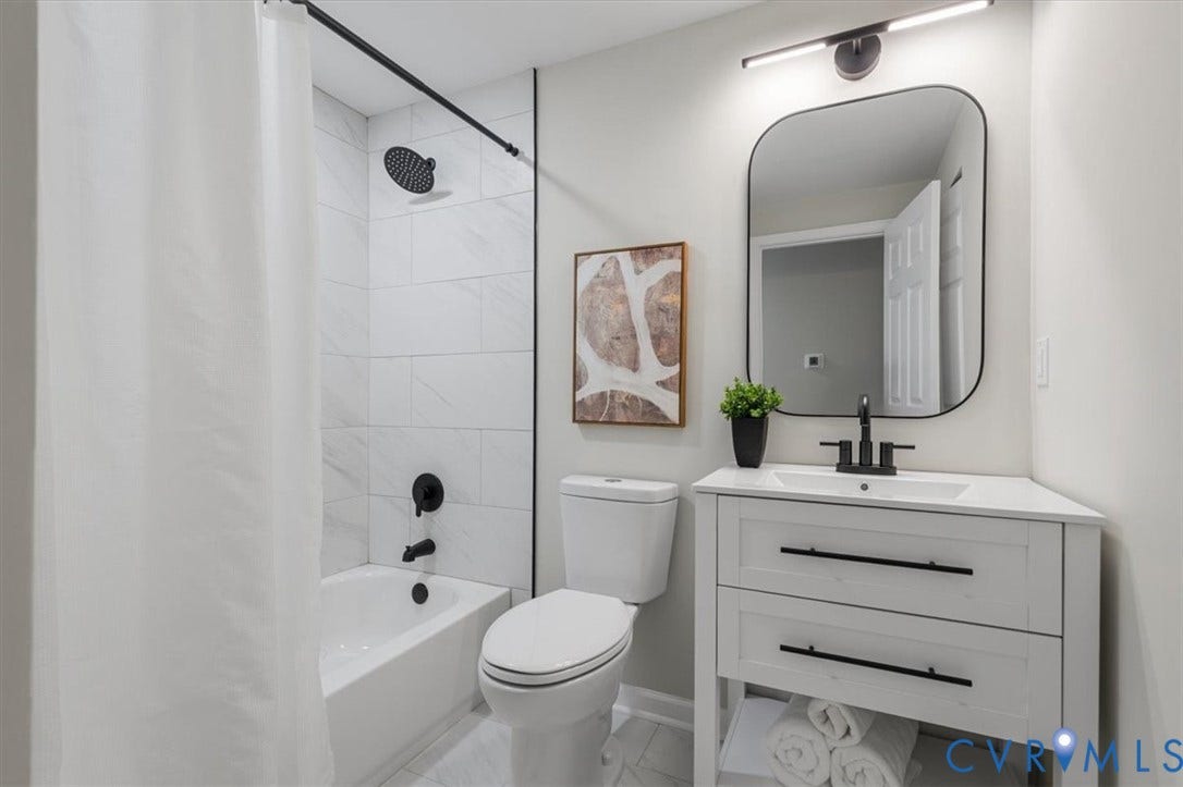 9201 Packard Court, Glen Allen, VA 23060 Bathroom featuring a white vanity with integrated rectangular sink, matte black hardware, stone-finish shower surround with rainfall showerhead, and a rectangular wall mirror with rounded corners