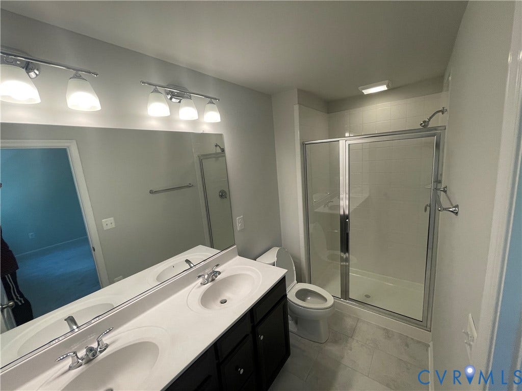 9212 St Charles Circle, Richmond, VA 23235 Dual-sink vanity with white countertops, dark cabinetry, full-width mirror, glass-enclosed shower, and large-format tile flooring