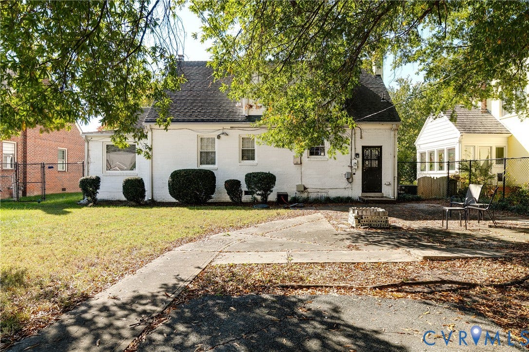 1406 Wentbridge Road, Richmond, VA 23227 Exterior featuring a painted brick facade, steep-pitched gable roof, concrete patio, mature trees, and perimeter chain-link fencing