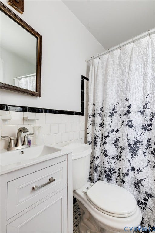1406 Wentbridge Road, Richmond, VA 23227 White vanity cabinet featuring a metallic finish faucet, wall-mounted mirror with a wood-tone frame, white wall tiling with black accent trim, and patterned tile flooring