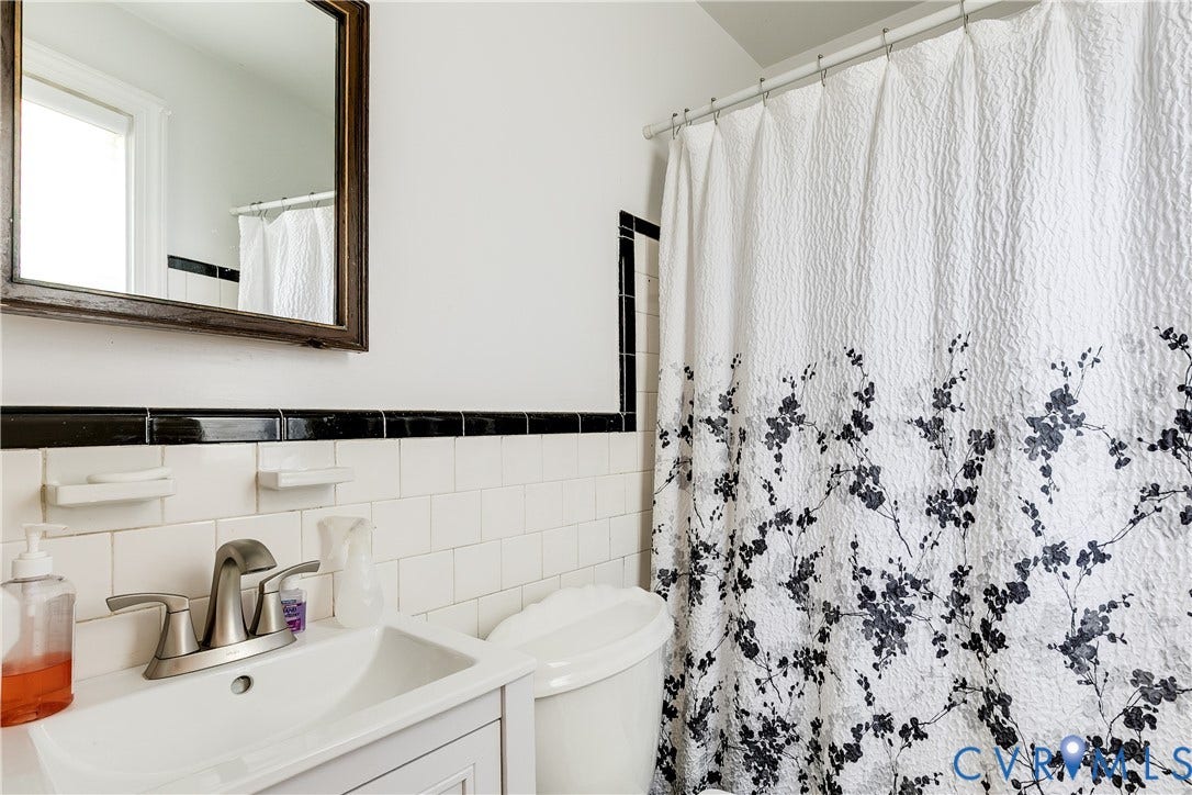 1406 Wentbridge Road, Richmond, VA 23227 Wall-mounted mirror with a dark wood frame, white ceramic square wall tile, decorative black accent tile border, porcelain sink, and satin-finish metal faucet