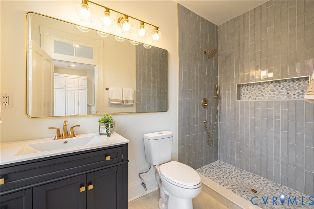 1108 W Marshall Street, Richmond, VA 23220 Dark-toned vanity with integrated white countertop, gold-finish faucet, wall-mounted light fixture, rectangular mirror, and walk-in shower with vertical tile surround and mosaic-tile niche