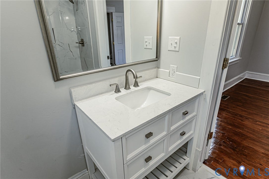 710 La Von Drive, Henrico, VA 23227 White vanity cabinet featuring a quartz countertop, an undermount rectangular sink, and brushed metal hardware, complemented by a silver-framed mirror and a wall-mounted faucet