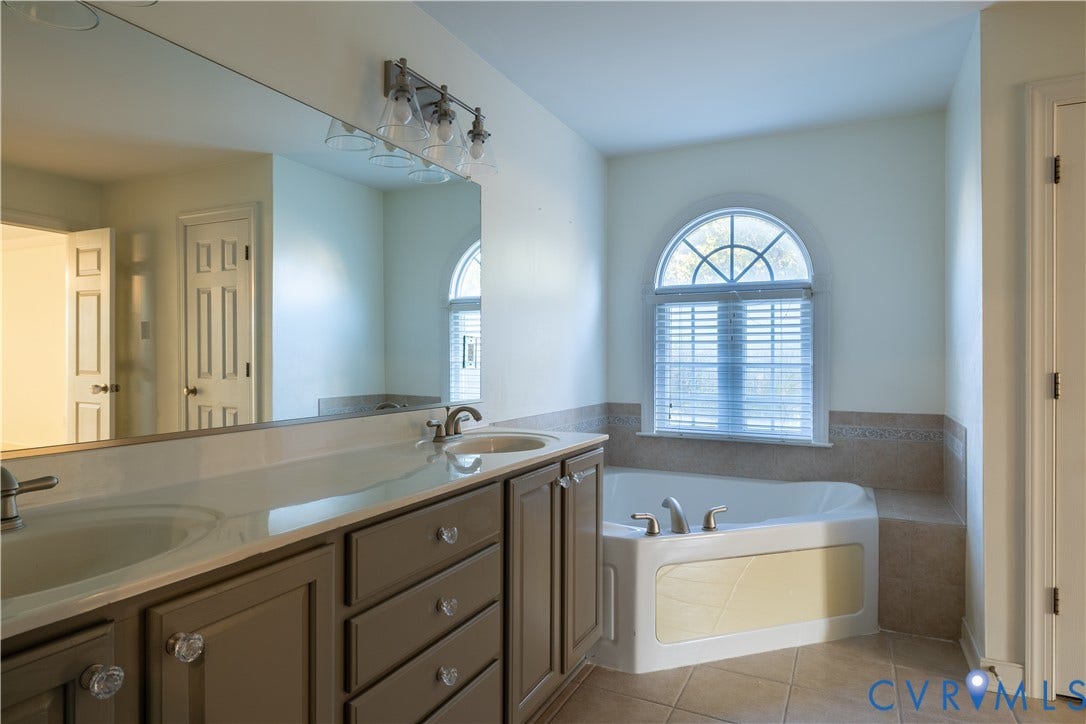Dual-sink vanity with neutral cabinetry, expansive wall-to-wall mirror, corner soaking tub, arched window, and ceramic tile flooring