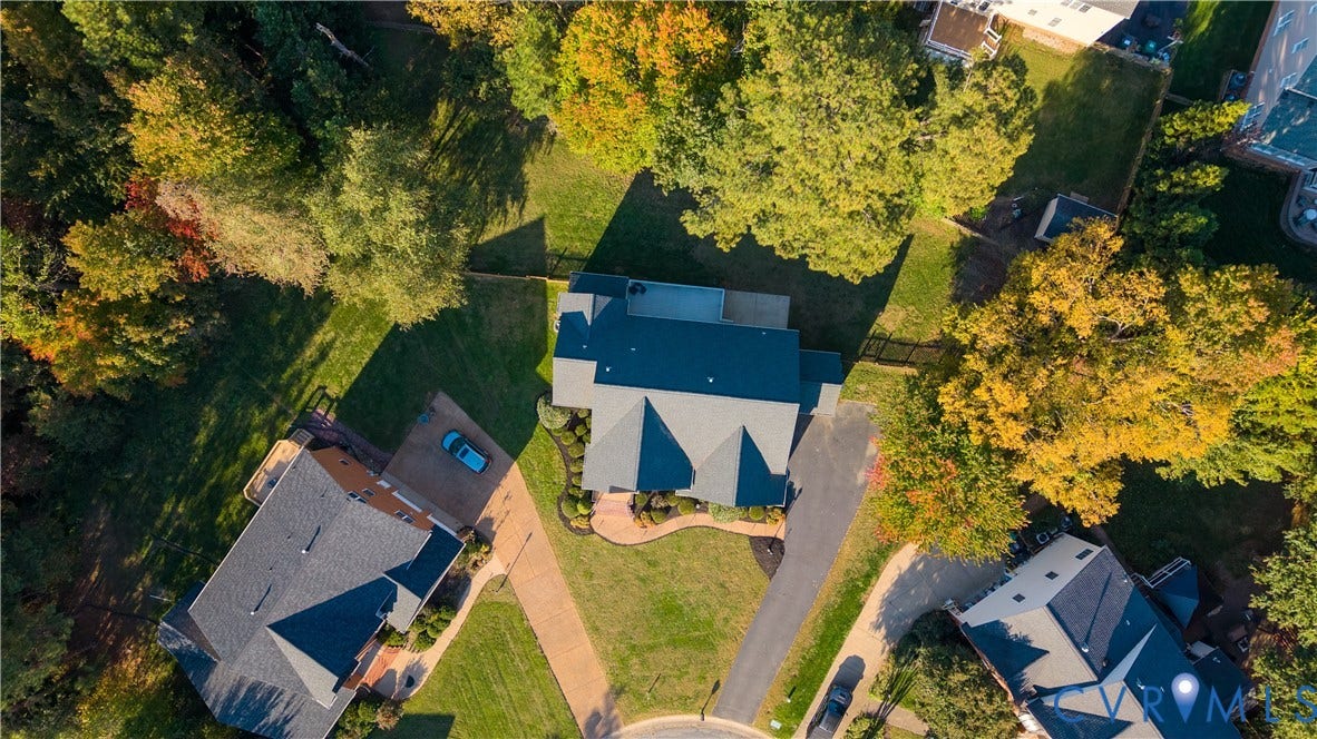 Aerial perspective featuring a multi-pitched shingled roof, asphalt driveway, paver walkway, and expansive lot with mature trees