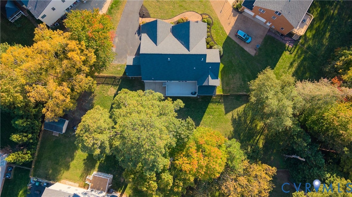 Aerial perspective featuring a multi-pitched asphalt shingle roof, a rear deck, a fenced perimeter, a paved driveway, and mature trees on the lot