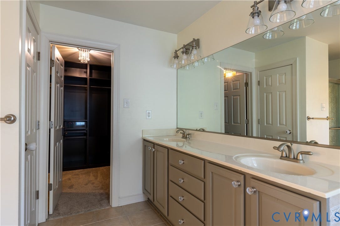 Dual-sink vanity with light-tone cabinetry, expansive wall-to-wall mirror, polished metal fixtures, and integrated storage, featuring a recessed walk-in closet with custom built-in shelving