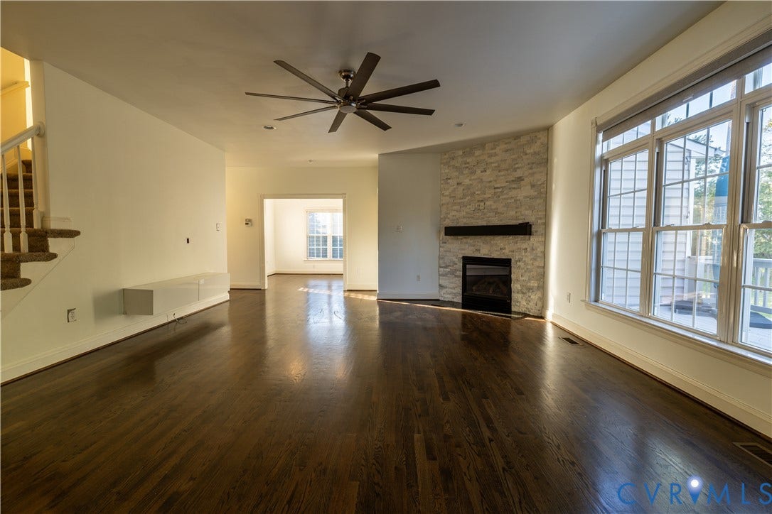 Living area featuring wood-finish flooring, stone-surround corner fireplace with a linear mantle, large multi-pane windows, and a ceiling fan