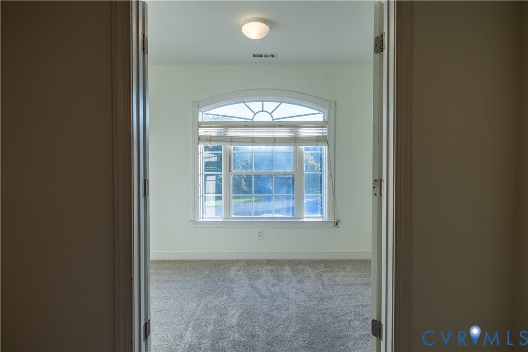 Double French doors opening to a room featuring neutral carpeting, a large window with a semicircular transom, and a flush-mount ceiling light