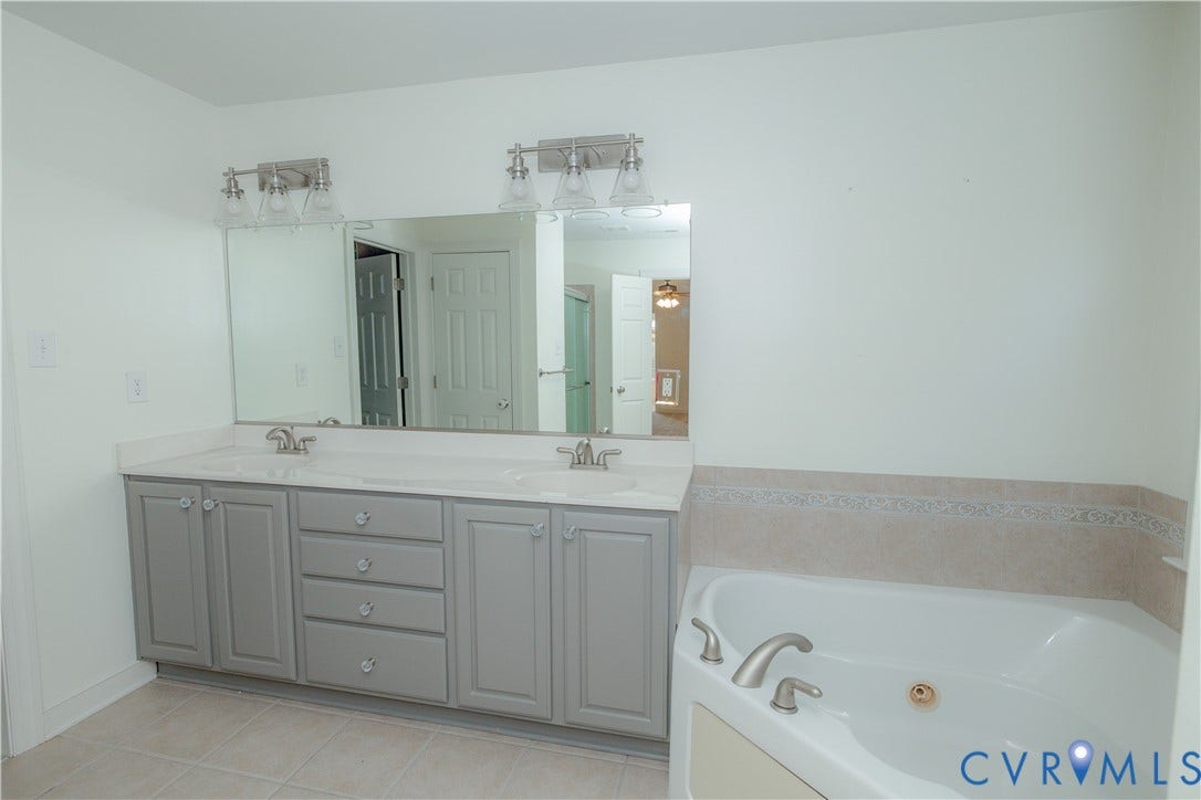 Dual-sink vanity with light gray cabinetry and integrated countertop, expansive wall-mounted mirror, brushed-nickel lighting fixtures, and a soaking tub with a tiled surround