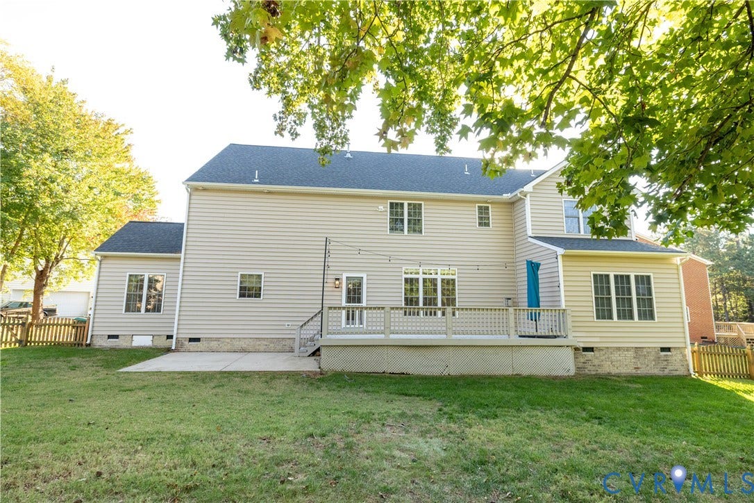 Exterior rear elevation featuring light-toned horizontal siding, a raised deck with lattice skirting, an integrated concrete patio, and mature trees