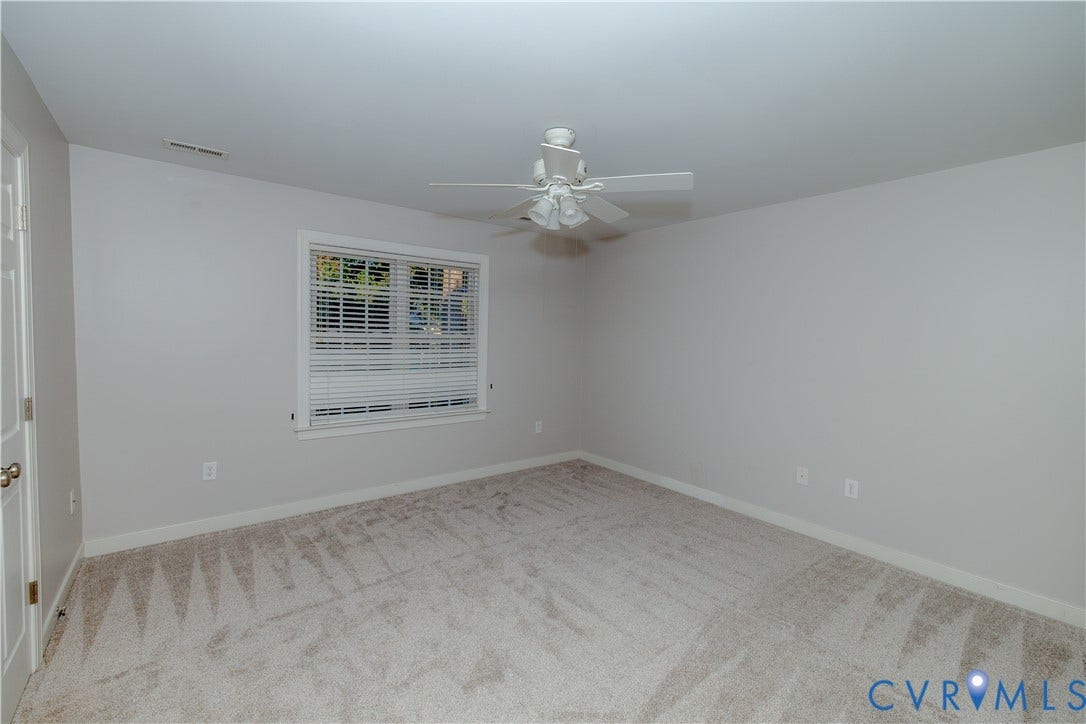 Neutral-toned interior featuring wall-to-wall carpeting, a multi-blade ceiling fan with integrated lighting, and a window with horizontal blinds