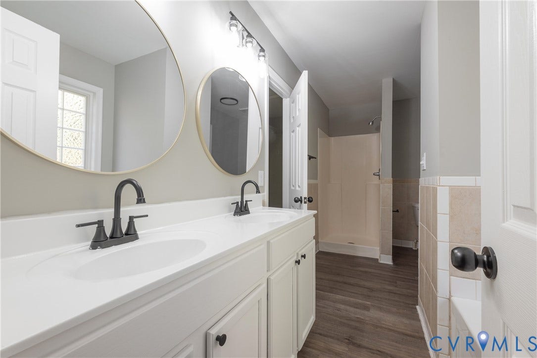 7009 Summers Trace Court, Chesterfield, VA 23832 Dual-sink vanity with white cabinetry, light-toned countertop, matte black fixtures, circular wall-mounted mirrors, and wood-finish flooring