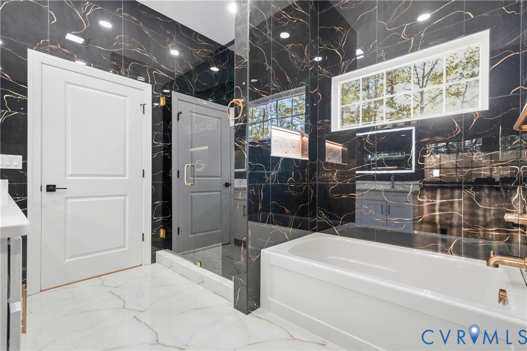 Bathroom featuring black wall tile with copper-toned veining, deep soaking tub, glass-enclosed shower, transom window, and white tile flooring with veining