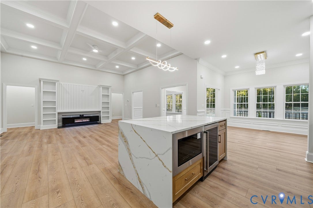 Coffered ceilings, center island with stone waterfall edges, built-in beverage refrigerator, wood-finish flooring, and integrated wall shelving with a built-in fireplace