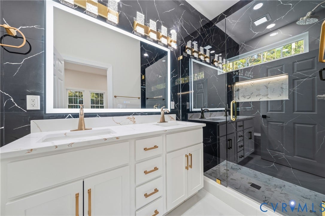 Dual-vanity bathroom featuring large-format dark veined tile, white cabinetry with gold-toned hardware, solid-surface countertops, recessed lighting, and a frameless glass-enclosed shower