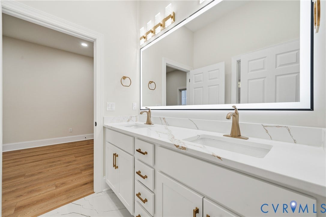 Dual-sink vanity with stone-look countertop, integrated rectangular basins, brushed gold-finish fixtures, oversized LED-backlit mirror, and white shaker-style cabinetry with matching gold-finish hardware