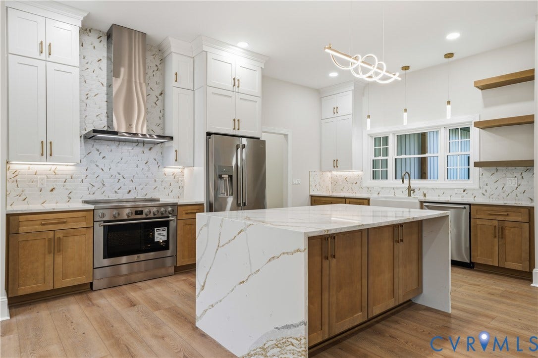 Kitchen featuring an oversized island with a waterfall countertop, two-tone cabinetry, patterned tile backsplash, stainless steel appliances, and floating wood-finish shelves
