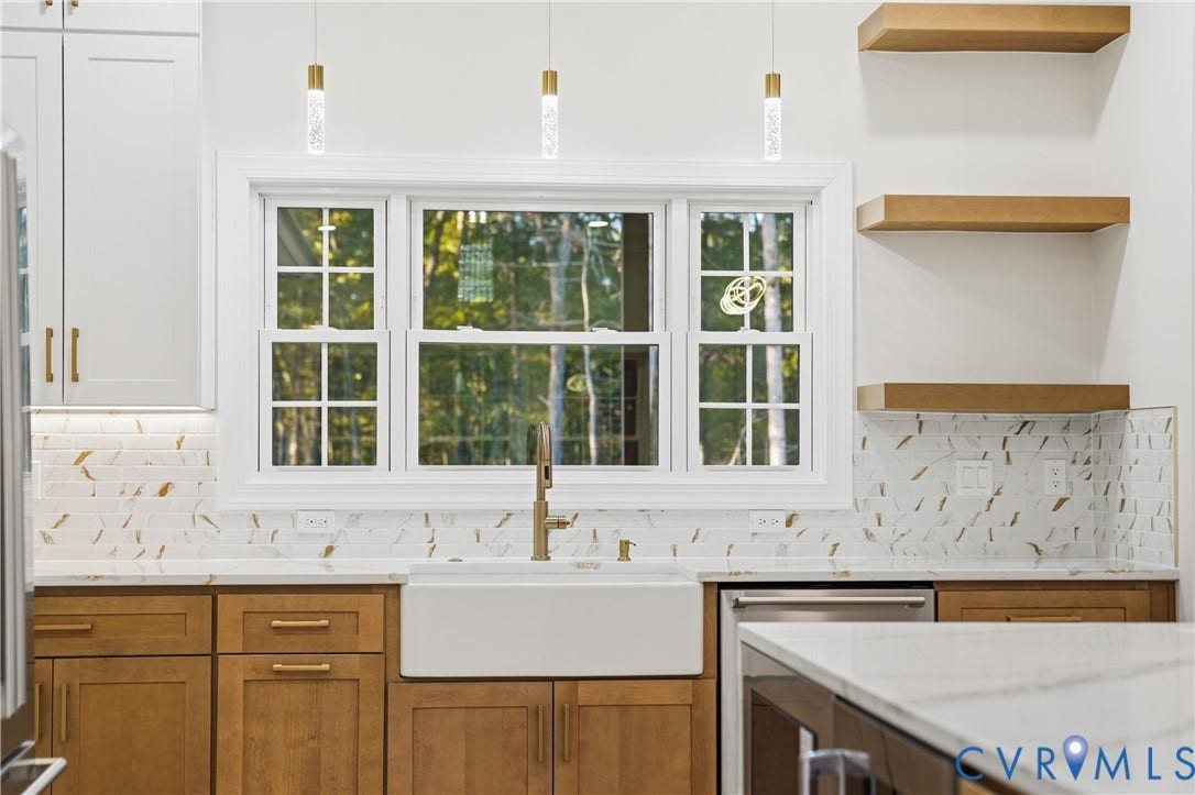 Kitchen featuring wood-finish cabinetry, white farmhouse apron sink, metallic-finish hardware, patterned tile backsplash, and floating wood shelving