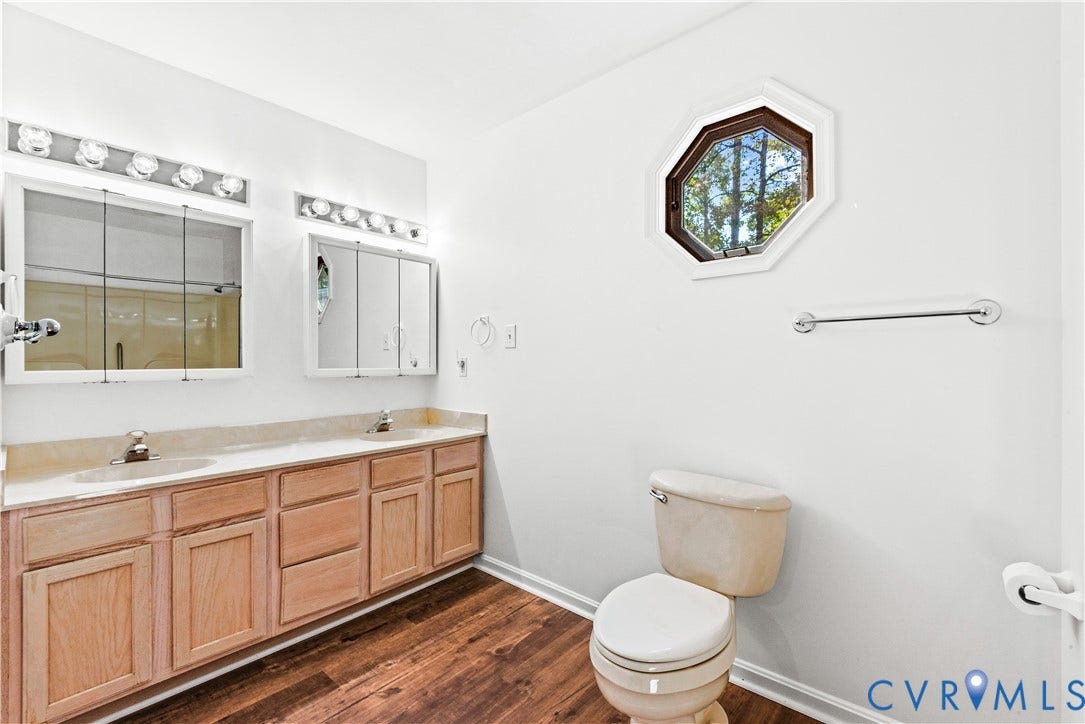 20040 Paradise Lane, Ruther Glen, VA 22546 Double vanity featuring wood-finish cabinetry, integrated dual sinks, mirrored medicine cabinets, and decorative wall lighting, complemented by wood-finish flooring and a geometric accent window