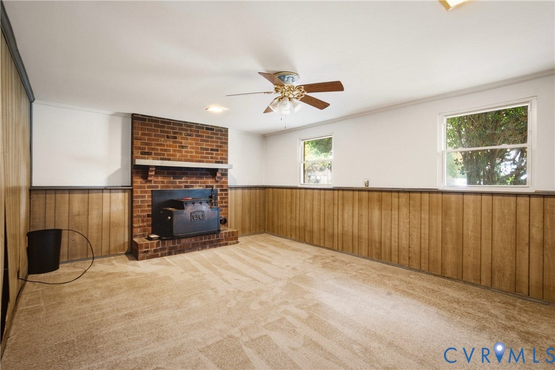 Living area featuring floor-to-ceiling wood paneling, a full-height brick fireplace with a wood-burning insert, wall-to-wall carpeting, and a ceiling fan with integrated lighting