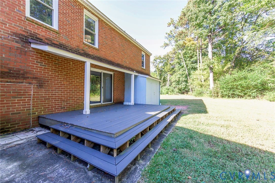 4124 Cogbill Road, Ampthill, VA 23234 Multi-level rear deck featuring wood-finish decking, structural support columns, a sliding glass door, and exterior brick cladding, set against a tree-lined backyard