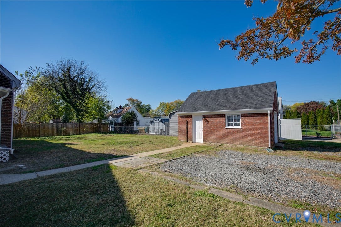 24 N Elm Avenue, Henrico, VA 23075 Detached brick structure featuring a gable roof, exterior lighting, and a gravel parking area, complemented by a grass yard and wooden privacy fencing