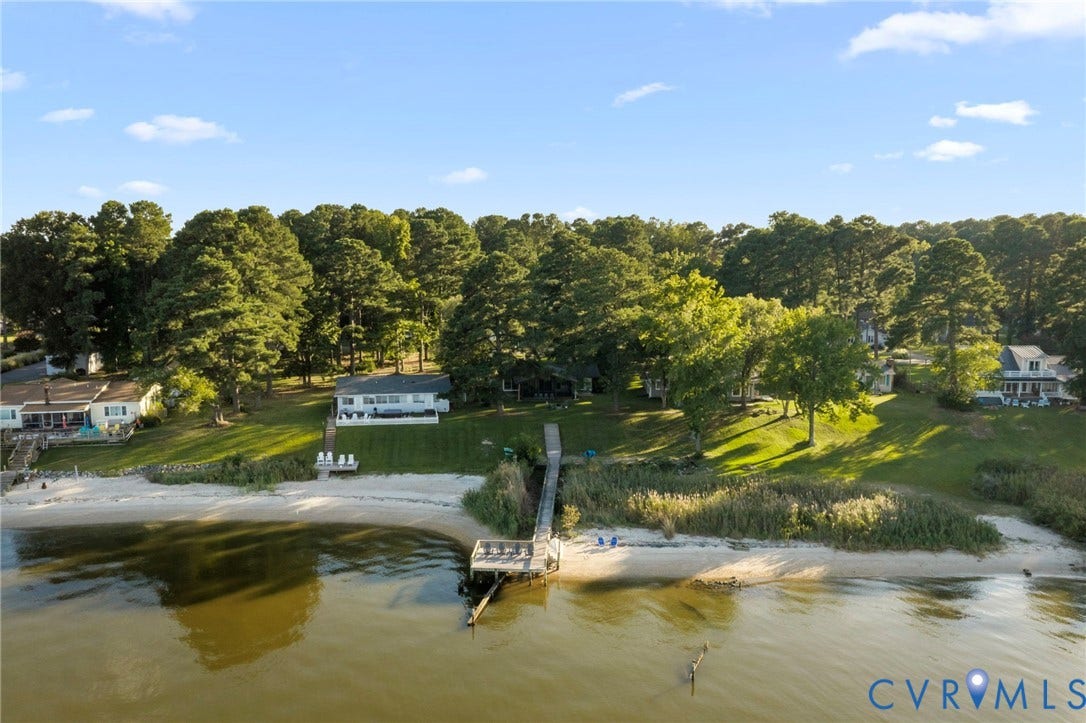 Waterfront property featuring a private wooden pier, expansive sandy beach area, mature trees, and tiered lawn space