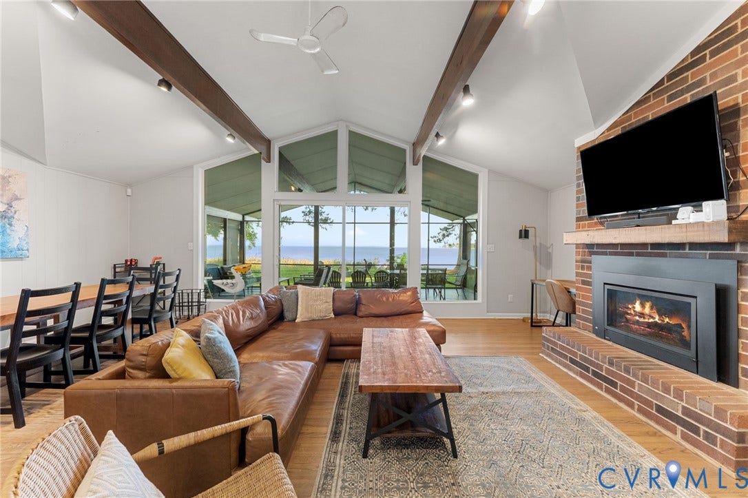 Vaulted ceilings with exposed wooden beams, brick fireplace with a custom wood mantel, wood-finish flooring, and expansive floor-to-ceiling windows