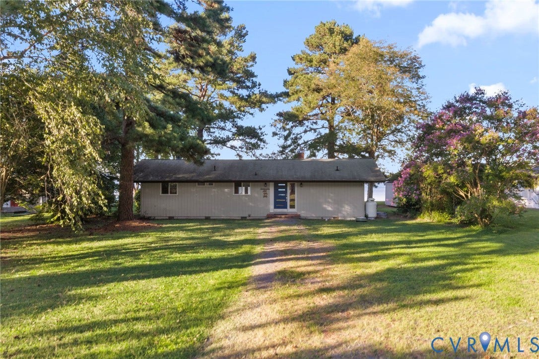 Single-story residence featuring light-toned horizontal siding, a covered entryway, and a low-pitched roof, set upon a spacious lot with mature trees