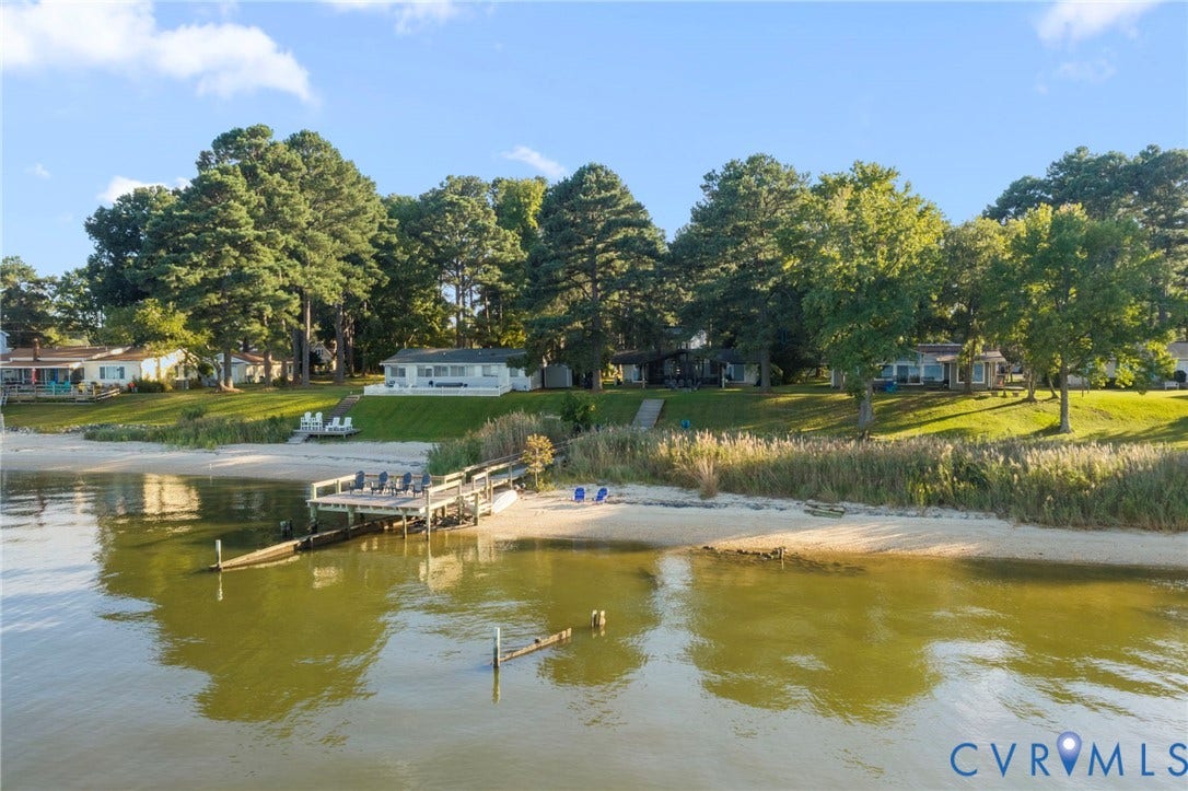 Waterfront property featuring a private wooden pier, sandy shoreline, grass-covered grounds, and mature trees