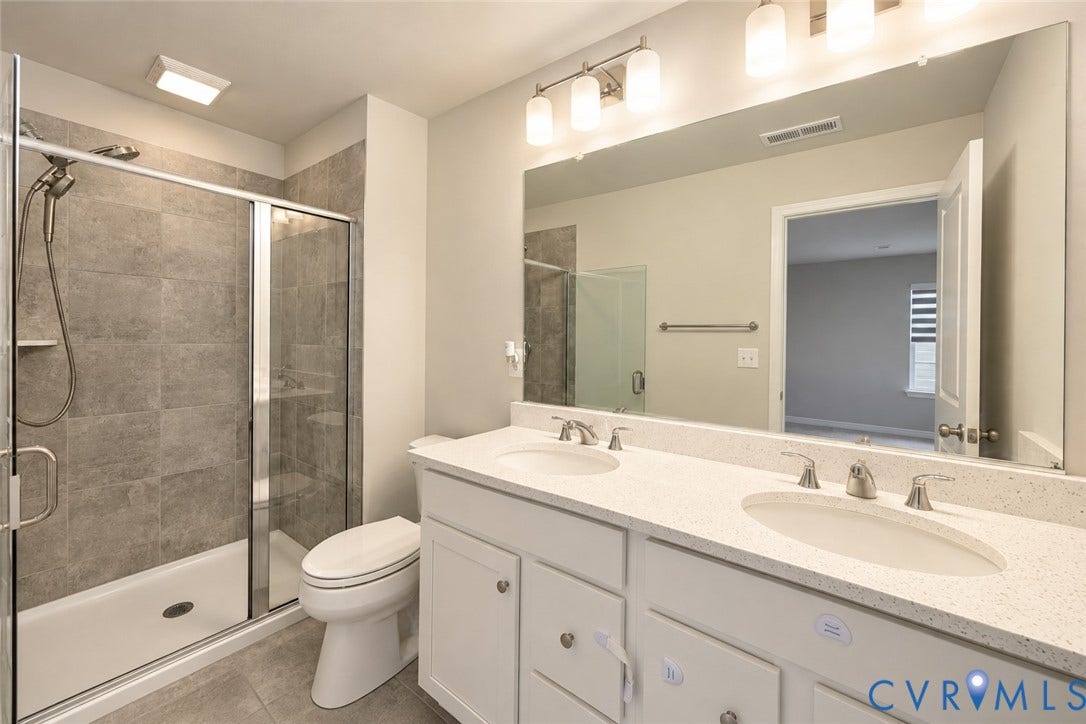 500 Rivanna Hill Way, Glen Allen, VA 23060 Bathroom featuring a dual-sink vanity with a solid-surface countertop, full-width mirror, glass-enclosed shower with tiled surround, and multi-light fixtures