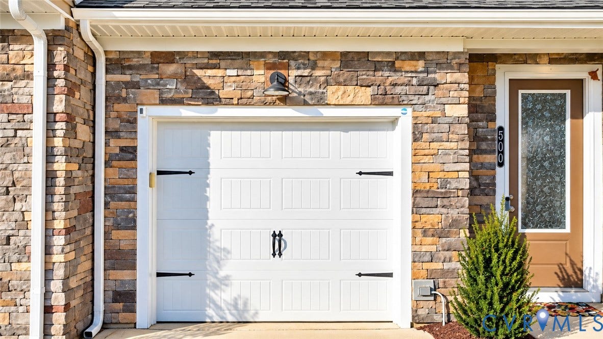 500 Rivanna Hill Way, Glen Allen, VA 23060 Multi-textured stone facade featuring a white carriage-style garage door with decorative black hardware, an exterior wall-mounted gooseneck light fixture, and a glass-paneled entry door