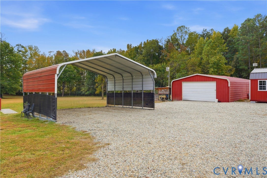 16401 Anderson Drive, Disputanta, VA 23833 Metal-roofed carport featuring a gravel-surfaced drive, red steel-sided outbuilding with a roll-up garage door, and a wooded perimeter